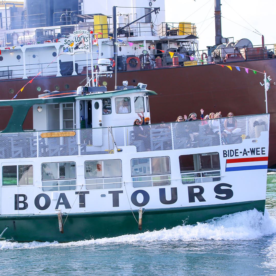 Passengers on the Original Soo Locks Tours wave to the camera as a freighter passes behind them.