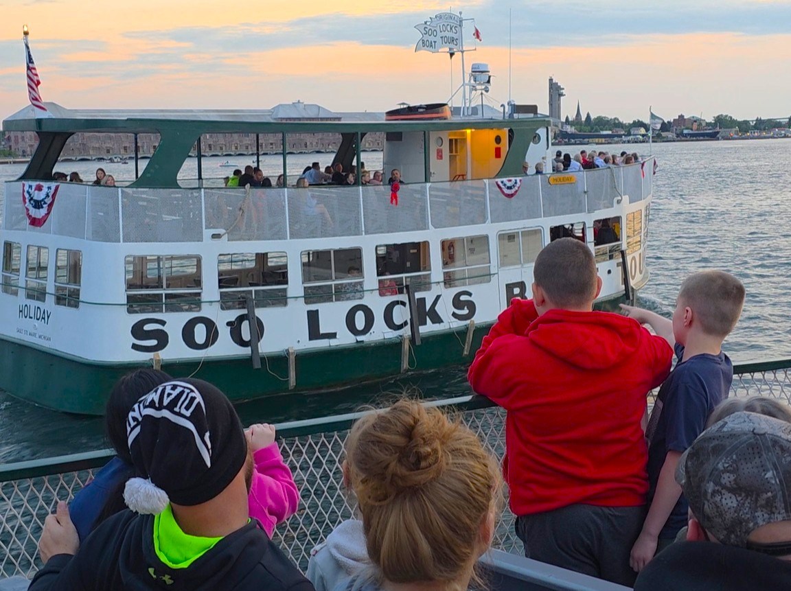 Kids on deck of the Original Soo Locks Tours Boats making their way to the Soo Locks with a beautiful sunset in the foreground
